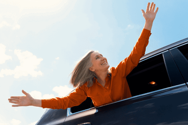 A person in an orange shirt leans out of a car window with arms outstretched, smiling against a blue sky background.