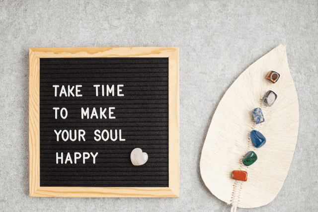 A wooden-framed board displays the text, "Take time to make your soul happy." Next to it, a decorative leaf holds six colorful stones.