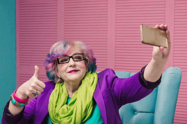 An elderly person with colorful hair takes a selfie while giving a thumbs-up. They wear glasses, a purple jacket, and a bright green scarf, sitting on a teal chair against a pink background.