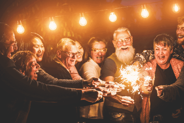 A group of people smiling and holding sparklers under string lights during a nighttime gathering.