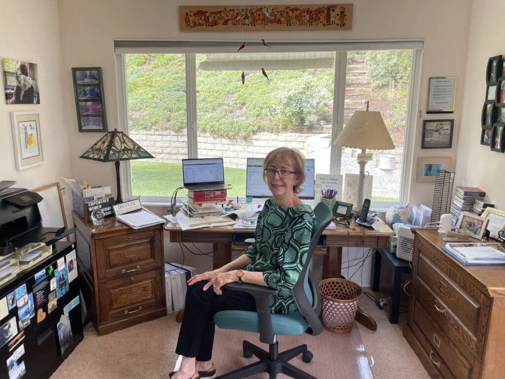 A woman sits in a home office with a large window behind her. The office is furnished with a desk, a computer, and various office supplies.