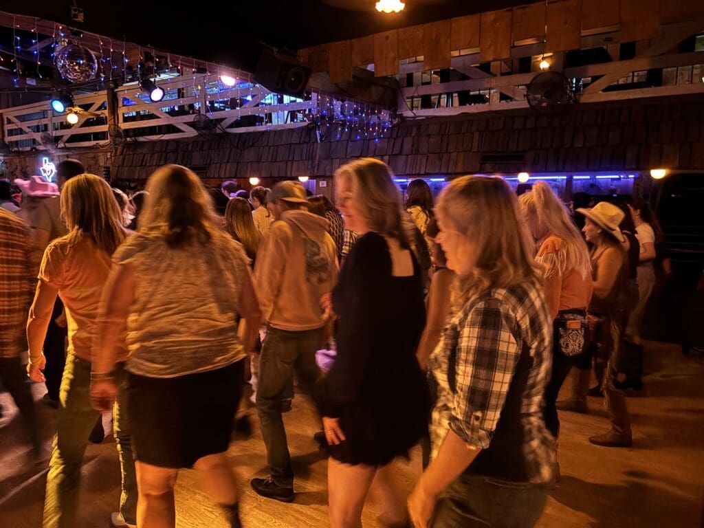In a dimly lit venue with rustic wooden decor, people are rhythmically line dancing, their movements synchronized to the lively music.