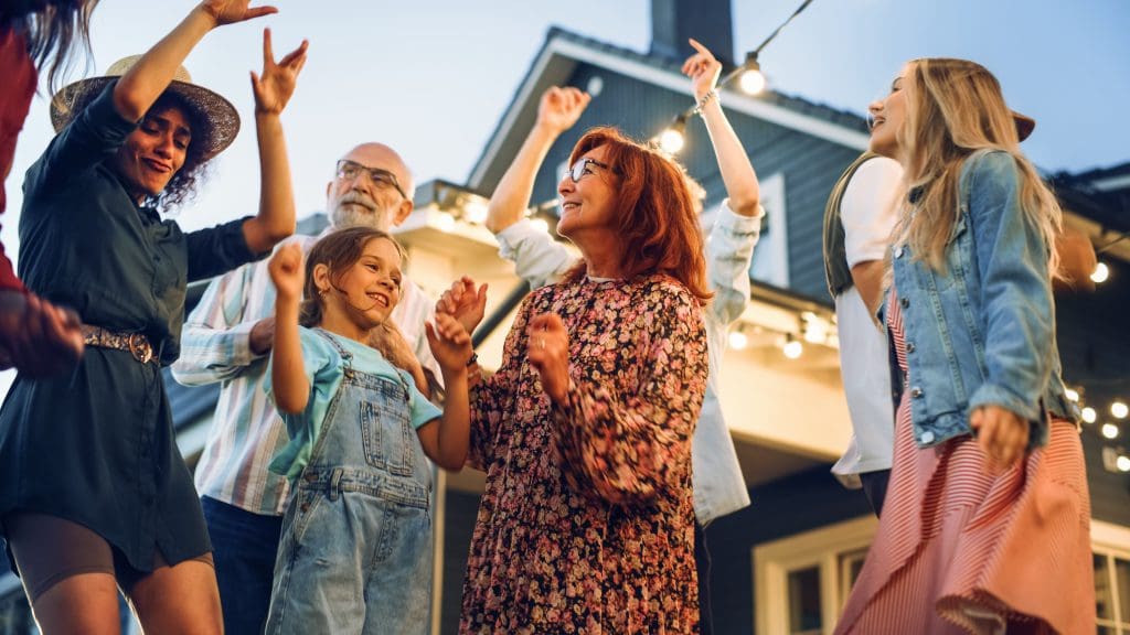 A group of people, including adults and children, are dancing happily outside near a house, under string lights, during the evening.