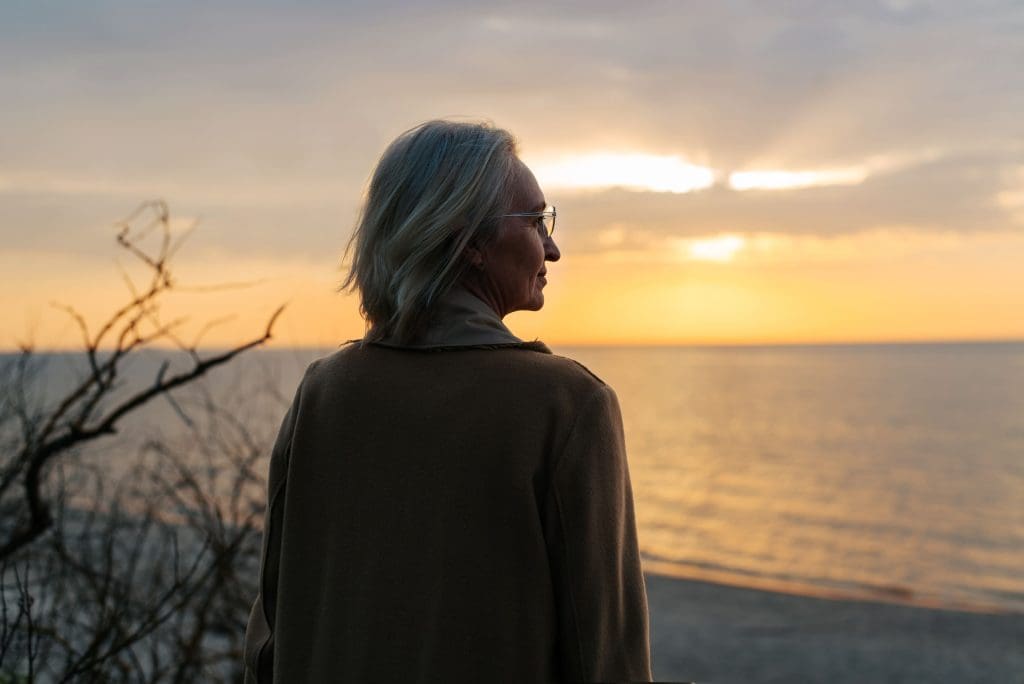 A person with shoulder-length hair and glasses stands on a beach, watching the ocean and sunset. There are branches in the foreground.