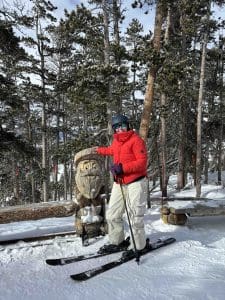 A person in a red jacket and ski gear stands on skis in the snow, posing next to a wooden carving of a bearded face in a forested area.