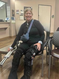 A woman wearing black clothing sits in a waiting room with both legs in orthopedic braces, holding a crutch and carrying a small blue bag across her chest.