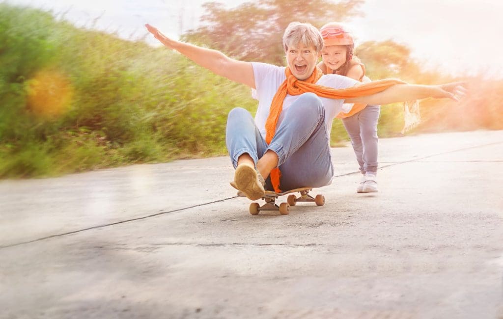 Older woman sits on a skateboard with arms outstretched, smiling, while a young girl wearing a helmet pushes her from behind on a sunny outdoor path.