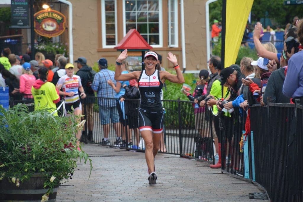 A triathlete smiles and raises her hands while running past cheering spectators during a race in an outdoor setting.
