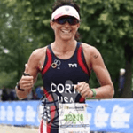A triathlete wearing a USA uniform and sunglasses runs outdoors, smiling and giving two thumbs up, with trees and event barriers in the background.