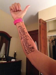 A persons forearm with a handwritten list of names and numbers in black marker, wearing a pink watch, with a raised thumb in a bedroom setting.