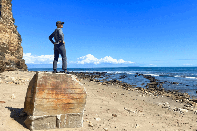 Person standing on a large rock on a sandy beach, facing the ocean, with cliffs and blue sky in the background.