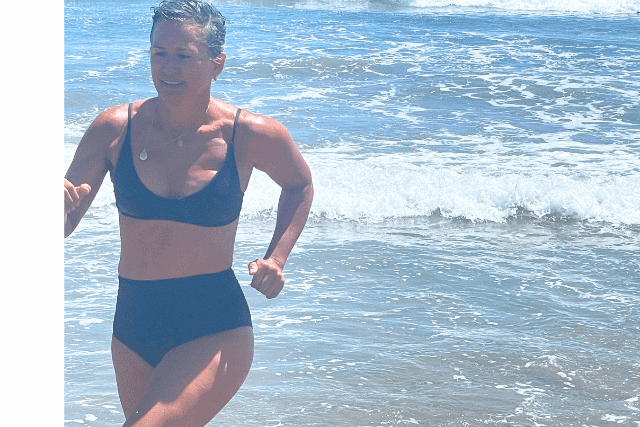 A person in a black swimsuit runs along the shore with ocean waves in the background.