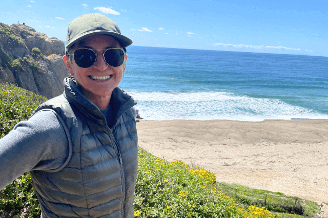Person wearing sunglasses, a cap, and a puffy vest smiles while standing on a coastal cliff overlooking a sandy beach and the ocean on a sunny day.