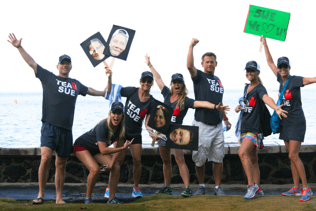 A group of seven people wearing matching TEAM SUE shirts pose enthusiastically by the water, holding signs with faces and a green poster that reads SUE WE ♥ U!.