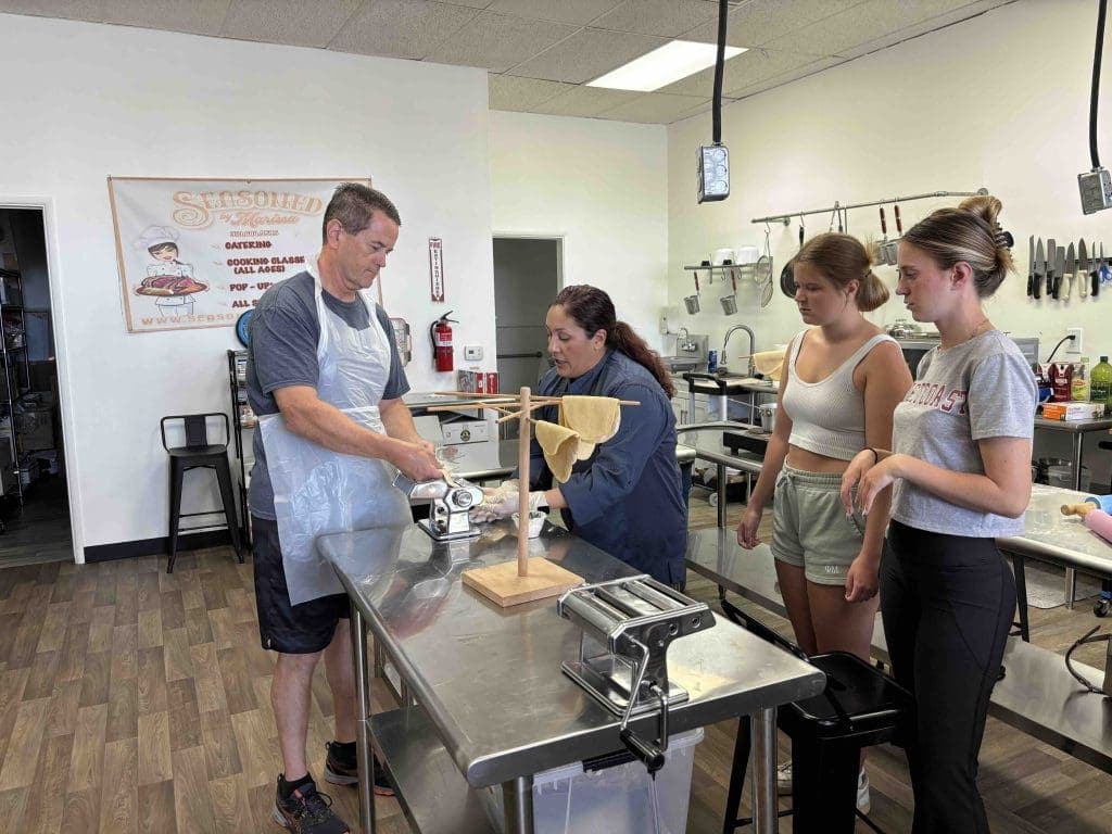 Four people in a kitchen watch as a man and a woman use a pasta machine; fresh pasta sheets hang on a wooden rack nearby.