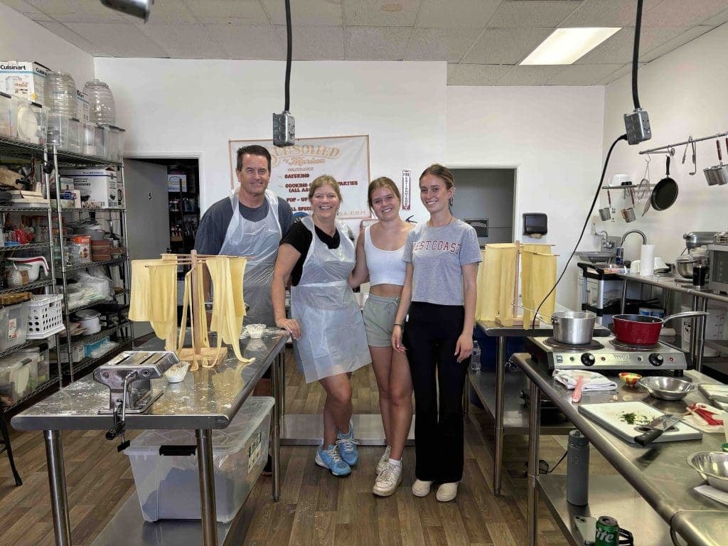 Four people stand together and smile in a commercial kitchen with pasta dough hanging to dry on racks and various cooking utensils and ingredients on the counters around them.