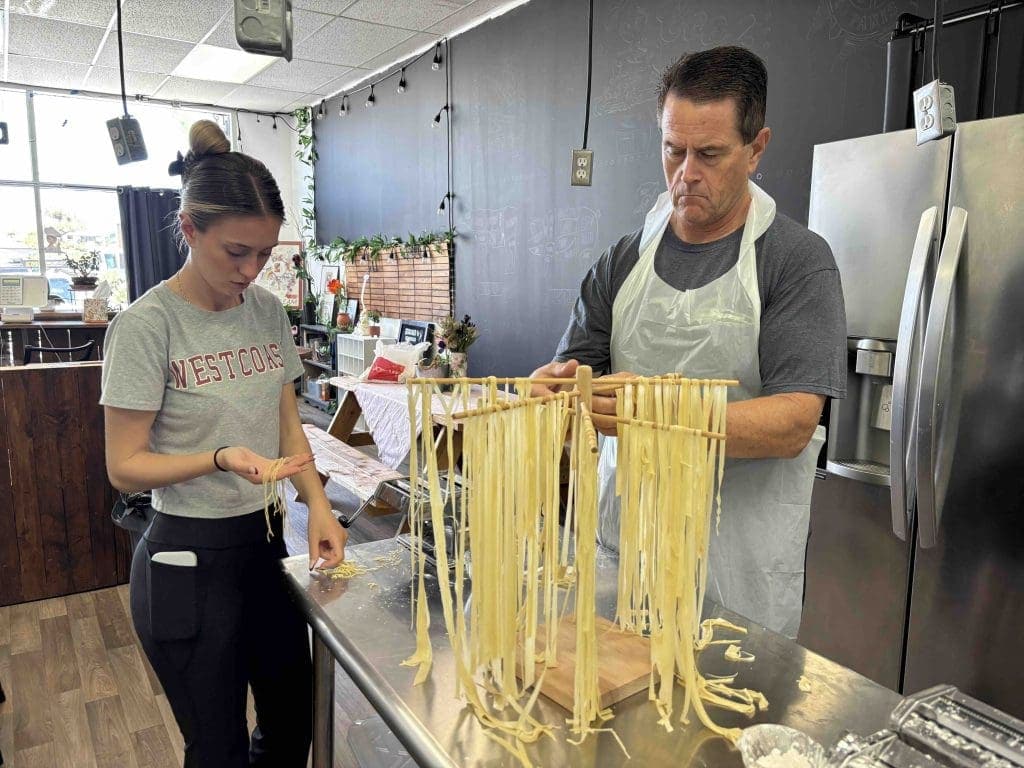 A man hangs fresh pasta on a wooden rack while a woman checks her hands in a kitchen setting.