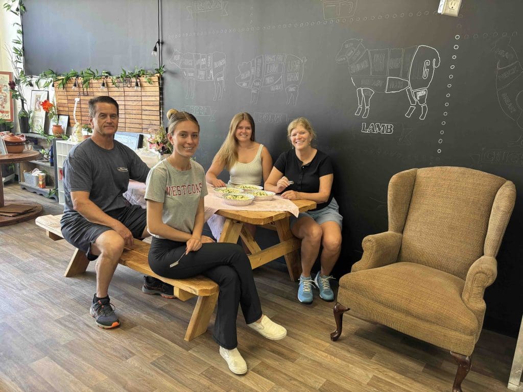 Four people sit around a wooden table with food inside a cafe. Behind them, a blackboard displays chalk drawings of animal meat cuts. A beige armchair is positioned to the right.