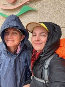 Two people wearing rain jackets and hats stand close together, smiling at the camera in front of a textured wall with a partial design visible behind them.