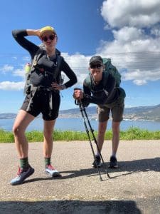 Two hikers with backpacks and hiking poles pose playfully on a paved path, with a scenic landscape, water, and mountains in the background under a partly cloudy sky.