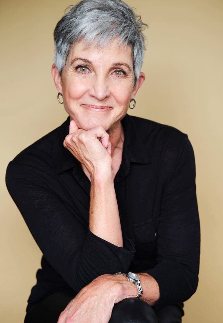 Older woman with short gray hair, wearing a black shirt and silver hoop earrings, sits smiling with her chin resting on her hand against a beige background.
