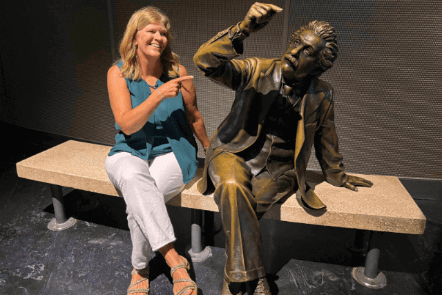 A woman sits on a bench next to a bronze statue of Albert Einstein, both pointing at each other and smiling.