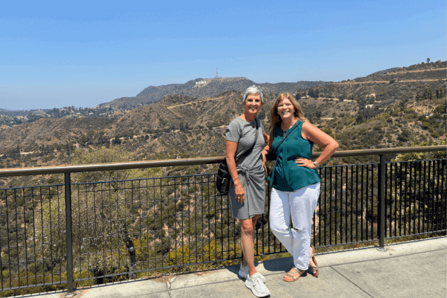 Two women posing and smiling in front of a railing, with hills and the Hollywood Sign visible in the background on a clear day.