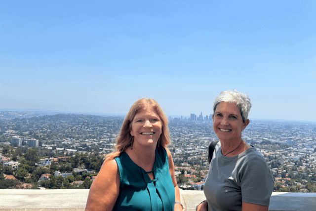 Two women stand in front of a cityscape on a clear day, with a distant skyline and buildings visible under a blue sky.