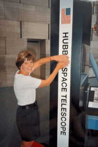 A woman stands next to a vertical Hubble Space Telescope sign indoors, smiling and placing her hands on the sign.