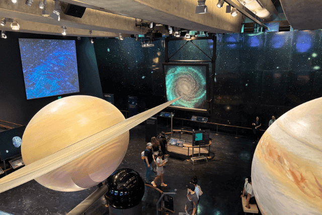 People observe large models of planets, including Saturn and Jupiter, in a space-themed museum exhibit with cosmic backgrounds and interactive displays.