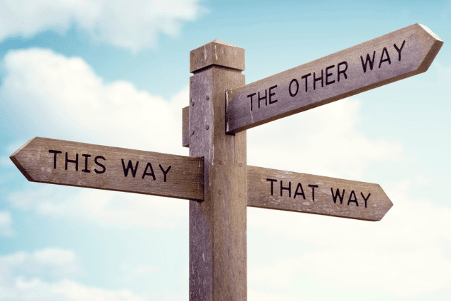 A wooden signpost with three arrows labeled THIS WAY, THE OTHER WAY, and THAT WAY against a blue sky with clouds.