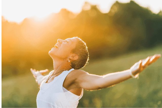 A person standing outdoors with arms outstretched, facing the sunlight, wearing a white sleeveless top and smiling, with a blurred natural background.