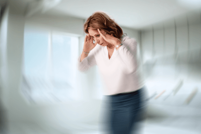 A woman holds her head with both hands, appearing distressed, with the image blurred around her to suggest dizziness or confusion.