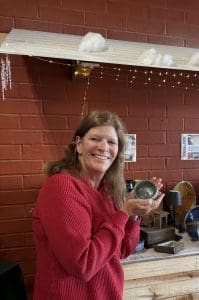 A woman in a red sweater smiles and holds a small round object in both hands, standing in front of a red brick wall with shelves and decorative lights.