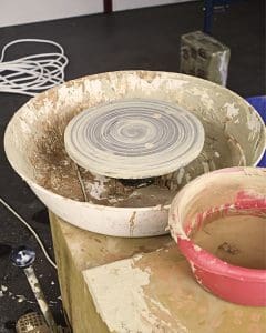 A pottery wheel covered in clay sits next to a red bowl filled with wet clay, with a messy workspace and scattered tools in the background.
