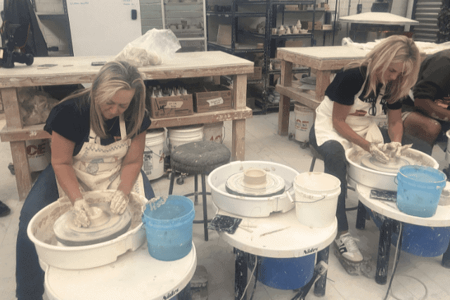 Two women seated at pottery wheels in a studio work with clay, each shaping pottery on separate wheels with tools and supplies nearby.