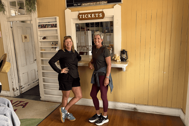Two women smiling at the ticket counter in a fitness or wellness center.
