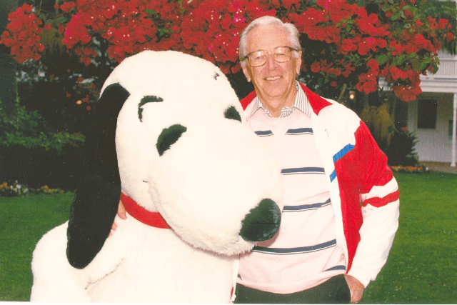 Charles M. Schulz with a Snoopy mascot outdoors in front of red flowers.
