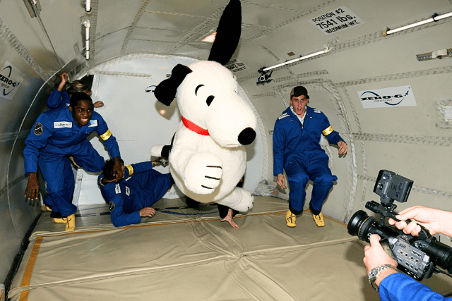 Astronauts and Snoopy mascot inside spacecraft for space training or event.